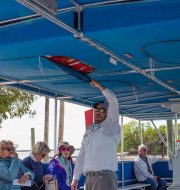 Tour guide standing out boat showing passengers the life vests storage above their heads